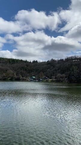 The video shows a view of a lake on a partly sunny day. The water is calm and reflects the sky. The opposite shore is lined with trees, and a few houses can be seen among the trees. The sky is blue with white, puffy clouds. The camera pans slowly to the right, revealing more of the lake and the shoreline. The overall impression is one of peacefulness and natural beauty.