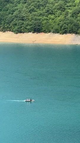 A white boat with people on board travels across a wide body of turquoise water. The water is calm with small ripples. In the background, a sandy shore meets a steep, tree-covered hillside. The trees are lush and green, indicating a warm season. The overall scene is serene and natural.