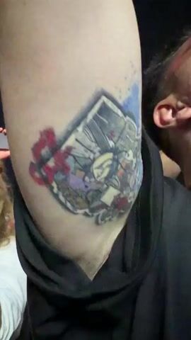The video opens with a close-up shot of a colorful tattoo on someone's upper arm. The tattoo appears to be a complex design with multiple figures and abstract shapes. The camera then pans to the right, revealing the face of a person holding up a smartphone, as if recording. The background is dark, with some blurred foliage visible. The scene then shifts to a wider shot of a concert or event. A stage with bright blue lights and a large screen displaying a figure can be seen. The word "PARK LIVE" is illuminated in orange letters above the stage. A large crowd of people is visible in the foreground, many holding up their phones to record. The lighting is predominantly blue and purple, with spotlights illuminating the stage. The overall impression is of a lively and energetic outdoor event.
