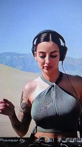 A woman is DJing in the desert. She has dark hair pulled back in a bun, headphones on, and is wearing a sleeveless gray top. She has tattoos on her right arm and hand. She is operating a black Pioneer DJ console. The background is a light tan desert landscape under a clear blue sky. She is looking at the equipment and occasionally up at the viewer, sometimes gesturing with her hands. There is music playing.