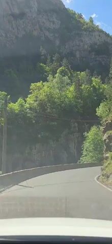 A car travels along a winding, paved mountain road on a bright, sunny day. The mountainside to the left is covered in lush, green trees and a large, rocky cliff face. A concrete barrier runs along the side of the road. Blue sky is visible above.