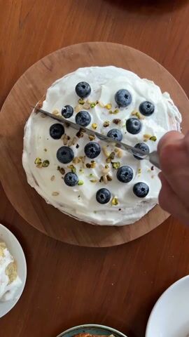 A person is cutting a cake with a small metal knife. The cake has white frosting and is decorated with blueberries, pine nuts, and pistachios. The cake sits on a round wooden plate. The table is brown wood. There is a partial slice of cake on a plate next to the cake.