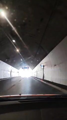 The video captures a first-person perspective from inside a moving vehicle, driving through a tunnel and then onto an open road. Inside the tunnel, artificial lighting illuminates the concrete walls and ceiling, while speed limit signs (one displaying "100" and another "80") are visible. The tunnel exit reveals a bright, sunlit landscape with a clear blue sky, fluffy white clouds, and distant mountains. The vehicle emerges onto a multi-lane highway, with streetlights lining the roadside and a rocky, barren mountain face visible on the right. No other vehicles or people are distinctly visible.