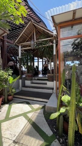 A woman walks up a short flight of stairs into a large, open-air restaurant. The restaurant has a thatched roof and a view of the ocean. There are plants in large pots on either side of the stairs. The woman is wearing a beige t-shirt, black pants, and sneakers. She smiles and waves at the camera as she walks up the stairs. A dog is lying down on the floor of the restaurant near the stairs.