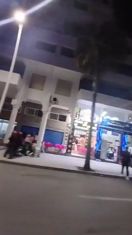 The video shows a night scene on a city street. On the right side of the street, there are white buildings with shops visible. A couple of cars pass by on the street. A small group of people are sitting on the steps of a building. The overall lighting is a mix of street lights and store illumination.