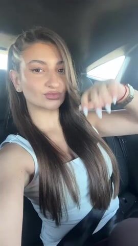 The video shows a woman in a car. She's wearing a white top and has brown hair. She poses for the camera, winking and smiling. She has a gold watch on her left wrist and a red string bracelet on her right. The interior of the car is visible, with black leather seats. The sun shines through the car window. The woman is wearing a seatbelt. There is music playing in the background.