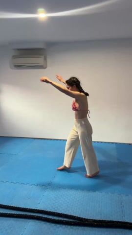 A young woman does a backbend in a brightly-lit room. She is wearing a pink bikini top and cream-colored linen pants. She is on a blue mat in a room that also contains an air conditioner and black ropes. She performs the backbend slowly, kicking her legs up and over her body.