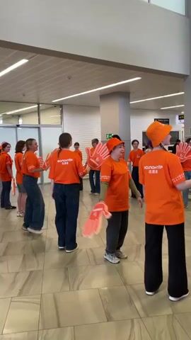 The video shows a lively scene at what appears to be an airport terminal. A group of people wearing bright orange shirts with "Volunteer" written in English and another language are standing around waving large, hand-shaped foam props, seemingly creating an enthusiastic welcome. 
Nearby, three women in traditional Russian folk costumes—featuring long, red skirts and blue blouses—are dancing and singing, adding a festive and culturally rich element to the atmosphere. The environment is a modern airport setting, with tiled floors, large pillars, and visible signage overhead. Passengers are seen moving around, some observing the performance.