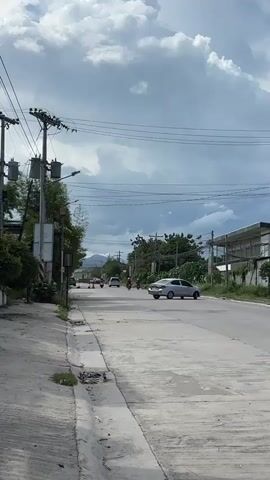 A vertical video captures a rural road with a cloudy sky overhead. On the left side of the road, a concrete sidewalk runs alongside a drainage ditch. The sidewalk is bordered by green foliage and utility poles with transformers. In the distance, several motorcycles and a silver car are visible on the road. The right side of the road features buildings and more greenery. The overall scene depicts a quiet, possibly developing, area.