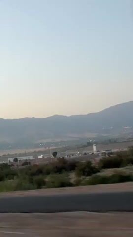 The video shows a blurred view from a moving vehicle looking out at a landscape. In the foreground, a blurred road barrier is visible. Beyond the barrier, there are trees and bushes. Further in the background, there are buildings, utility poles, and a road. In the distance, mountains form the horizon, and the sky is a pale, clear color. Several dark, indistinct shapes are visible in the sky above the landscape, appearing to be flying objects. The video quality is low, and the motion blur makes it difficult to discern fine details.