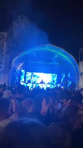 A vertical video shows an outdoor music concert at night. The crowd is visible from the waist up, facing a large stage with a domed roof. On the stage, a DJ is performing with mixing equipment. The stage is surrounded by powerful lights that change colors from blue to orange and red, illuminating the scene. Several pyrotechnic effects are seen throughout the video, including pillars of fire bursting upwards and smoke clouds. The crowd is energized, with some people dancing, raising their hands, and recording the event on their phones. The overall atmosphere is lively and festive.