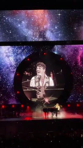 A concert performance features a male artist sitting at a grand piano on a stage. The artist is wearing a white shirt, a patterned vest, and a bandana. He is singing into a microphone, and a large screen behind him displays a close-up video of his face, also singing. The screen has a circular frame with red lights. The background of the large screen is a nebula with stars. Red smoke or fog is rising from the stage around the piano. A large audience is visible in the foreground, with their phones held up. The overall lighting is dark, with spotlights on the performer.