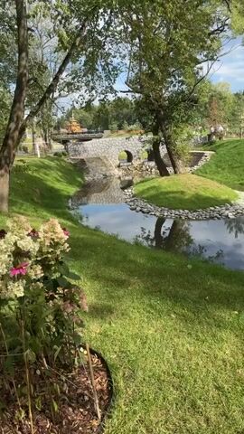 The video shows a serene park with a stream flowing under a stone bridge. The bridge has two arched openings through which water cascades down. Lush green grass lines the banks of the stream, with some potted flowering plants in the foreground. A tree stands on the right bank, casting a reflection in the water. In the background, a traditional wooden structure is visible beyond the bridge, and more trees form a natural backdrop. The scene is bathed in natural light, suggesting it is daytime. There are no people visible in the video.
