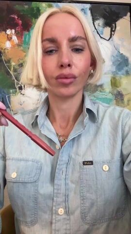 A fair-skinned woman with short blonde hair wearing a blue denim shirt with a Polo logo on the right side of her chest uses a CAIA Bryce-branded lip pencil to line her upper lip. She wears multiple bracelets and a gold ring. Tattoos are visible on the back of her hands. Behind her is a colorful abstract painting.