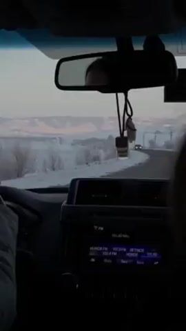 The video shows the interior of a car driving along a road in a winter landscape. The road is paved and there's snow-covered ground on either side. A few sparse trees can be seen in the snowy areas. In the distance, there are low-lying hills or mountains against a light pink and blue sky.

Inside the car, the dashboard and rearview mirror are visible. The rearview mirror reflects the face of the driver. A small decorative item hangs from the mirror. The car's radio display is visible, showing FM radio stations. Another car approaches from the opposite direction. The driver's hands are on the steering wheel.