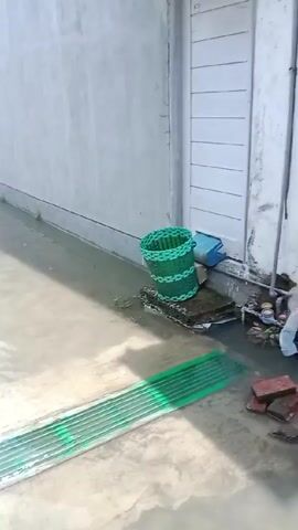 The video shows a flooded street. The water is high enough to partially submerge a yellow traffic gate. Two people are standing near a white building next to the gate. A motorcycle is visible in the distance.  The floodwater is murky.  The sky is blue. The environment appears to be a residential or commercial area.