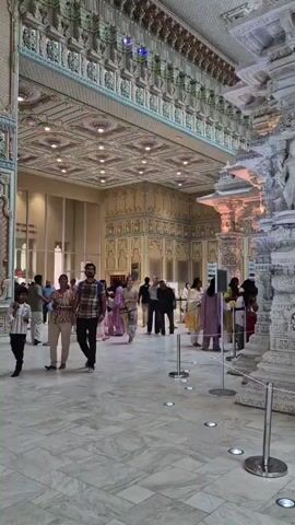 The video shows the interior of a large, ornate building, likely a temple. The architecture is elaborate, with intricately carved pillars, detailed ceilings, and decorative walls. The floor is marble. Several people are walking around the space, dressed in casual clothing, with some taking pictures. The overall atmosphere appears to be one of reverence and awe, with the focus on the detailed stonework and the grandeur of the building.