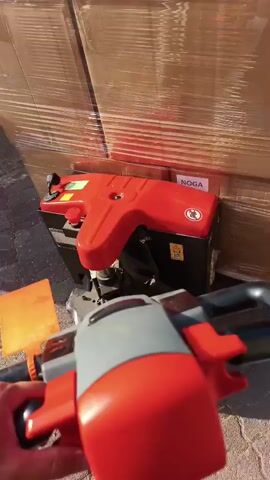 A first-person perspective video shows a person operating a red and grey pallet jack. The pallet jack is positioned in front of a large stack of brown cardboard boxes wrapped in clear plastic. The boxes appear to be on a wooden pallet. The pallet jack's handle and controls are visible in the foreground, and the lifting mechanism is extended under the boxes. The environment appears to be outdoors, with paving stones visible on the left side of the frame. The video is shot in daylight, and the lighting suggests a sunny day. There is a label on the boxes that reads "NOGA". The video is short, lasting only five seconds.