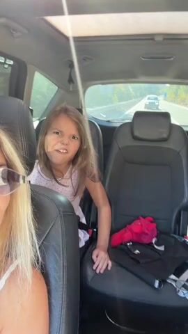 The video shows a young girl in the back seat of a car making faces at the camera. She is wearing a pink shirt and has her seatbelt on. In the front seat, a woman wearing sunglasses is also visible. The car is moving on a road, with other vehicles visible in the distance.