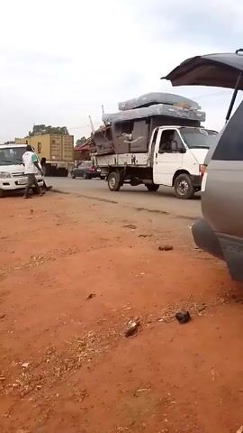 The video shows a roadside scene with several vehicles and individuals present. A white pickup truck is the focal point, with two men standing near its front. One man in a light-colored shirt appears to be working on the vehicle's front bumper or hood, while another man in a green shirt stands nearby. In the background, there is a road with passing vehicles, including a black sedan and a dark SUV with items on its roof. A truck loaded with furniture is also parked along the side of the road. The foreground consists of a reddish-brown dirt area with scattered debris. The sky is overcast, creating an overall muted lighting. There are also power lines and trees in the distance.