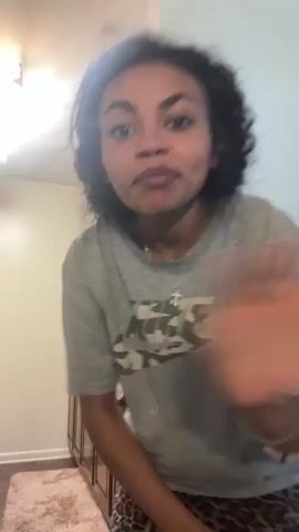 The video begins with a young woman with curly dark hair and a grey t-shirt that has a graphic print on it. She is smiling at the camera and wearing patterned pants. She moves closer to the camera, speaking directly to it. In the background, a younger person is visible sitting at a table in what appears to be a living room or dining area. The environment seems to be indoors with neutral-colored walls and a patterned wallpaper in the background. The lighting suggests it is daytime.