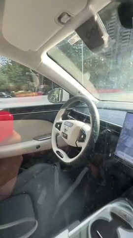 The camera is positioned inside a car, capturing the dashboard and the view through the windshield as the vehicle moves along a city street. The car's interior features a white and grey steering wheel with a logo in the center, a black dashboard, and grey seats. A dual-screen display is mounted on the center console, showing a real-time driving simulation or navigation interface. A dashcam is visible on the windshield. The outside environment, seen through the windshield, consists of traffic, buildings, trees, and street lights. The car appears to be equipped with advanced driver-assistance technology.