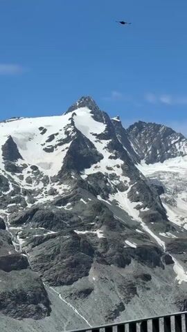 The video shows a panoramic view of snow-capped mountains against a clear blue sky. A bird can be seen flying high above the mountain peaks. The mountains are rugged and rocky, with glaciers and snow patches visible on their slopes. The camera pans downwards, revealing a pale green lake at the base of the mountains. In the foreground, part of a railing and some signage are visible, suggesting a viewpoint or observation deck. The overall scene is one of natural grandeur and a vast, untouched landscape.