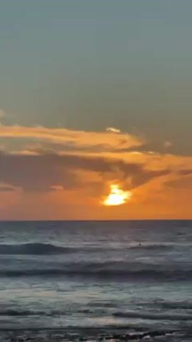 The video shows a sunset over the ocean. The sky is colored in shades of orange and grey, with the sun partially obscured by clouds. The ocean is dark and choppy, with small waves. There are a few figures visible in the water, presumably surfers. The foreground appears to be a rocky beach.