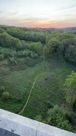 The video shows a high angle view of a bridge, a forest, and a field. The camera is initially focused on a path running through the field and up a hill. The sky is visible in the background, with colors of pink and orange, suggesting a sunrise or sunset. The camera pans upwards and towards the right, revealing the bridge structure and two men standing near the railing. The men are looking out at the scenery, one has his hand resting on the railing. They are both dressed in casual clothing. The environment is green and lush, indicating a natural, outdoor setting. The overall tone is peaceful and reflective.