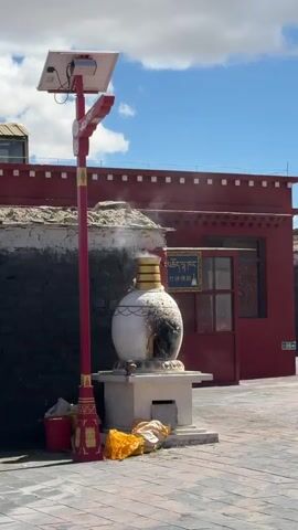 The video presents an outdoor scene in Labrang township, China, bathed in bright daylight under a clear blue sky with scattered white clouds. In the foreground, a prominent white, urn-shaped incense burner, adorned with a gold band at its neck and showing signs of charring from use, emits a continuous plume of white smoke. It stands on a white square base on a paved ground, with some yellow fabric or sacks resting nearby.

To the left of the burner is a tall red pole, likely a street light, topped with a small solar panel. In the background, a building with a vibrant red facade and a dark, traditional stone base is visible. A blue sign is affixed to the red building, featuring both Tibetan script and Chinese characters. The Chinese characters read "祈福增寿," translating to "pray for blessings and longevity," while the Tibetan script "བསང་མཆོད་དགོན་ཆེན་སྐྱེད" indicates "burning offerings to the great monastery." The active smoke from the burner suggests a ongoing ritual or ceremonial practice, such as burning sang (fragrant offerings), a common practice in Tibetan Buddhism for purification and making offerings. The scene evokes a strong sense of a spiritual or cultural site.