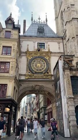 The video shows a street scene in front of a large, ornate clock mounted above an archway. People are walking in both directions on a cobblestone street. On the left, there are buildings with traditional architecture, including shops with awnings. On the right, a large banner is attached to scaffolding. The clock has Roman numerals and is gold and black. The sky is cloudy.