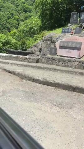 The video captures a memorial site nestled on a green hillside in Wazabaa, Abkhazia, under clear daylight. Lush green trees and dense foliage cover the steep slope in the background. In the foreground, concrete or stone steps ascend the hillside to the left, while a paved road or path occupies the very bottom of the frame, with a glimpse of a vehicle's interior in the bottom left corner.

The memorial itself comprises several dark, rectangular plaques mounted on a stone-faced structure built into the hillside. The central and largest plaque features black-and-white portraits of several men, with their names inscribed beneath them in Cyrillic, including "ШАЛАДЗЕ," "ЛАБАХУА," "АНЦУПОВ," "ШАРАШЕЛИЯ," and "ЛАГИЛАВА." Below these names, dates such as "1992" and "1993" are discernible, indicating the period of the Abkhazian War.

Above this central display, additional plaques are visible. One prominent plaque on the upper right side also shows portraits of individuals, accompanied by faint Cyrillic text that appears to mention "УЧАСТНИКИ АБХАЗСКОЙ ВОЙНЫ" (PARTICIPANTS OF THE ABKHAZIAN WAR). Smaller, individual memorial plaques with faces are also integrated into the stone work. The sunny weather illuminates the scene, highlighting the solemnity of the memorial against the vibrant natural backdrop. The overall setting is one of remembrance and honor for those commemorated.