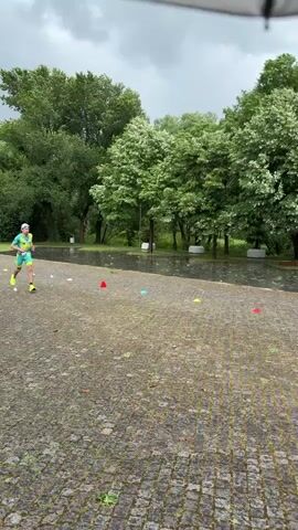 A man in a teal and yellow triathlete outfit with a white hat runs on a wet cobblestone surface in a park-like setting. He runs past several red cones and a red and black board lying on the ground next to a yellow container and a blue bag. Trees form a green backdrop, with some standing in water. The sky is overcast and it appears to be raining. The camera is static and offers a clear, steady view of the scene.