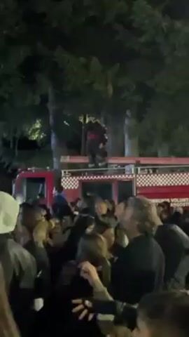 At night, a crowd of people is gathered around a red fire truck, where a person in a dark outfit with a red vest stands on top of the truck's roof. The camera zooms in on the person on the truck, then pans away towards a stage with a bright blue light shining on the performers, in front of another crowd of people. The stage is surrounded by tall, dark trees.
