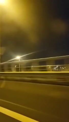 The video shows a view from a moving vehicle at night. The camera is looking out the side, capturing a highway or elevated road. There are streetlights illuminating the scene, creating streaks of light due to motion blur. The sky is dark, with some visible clouds. The road surface is visible with a yellow line marking the lane. A railing or barrier runs along the side of the elevated road. The overall impression is a nighttime journey on a road.