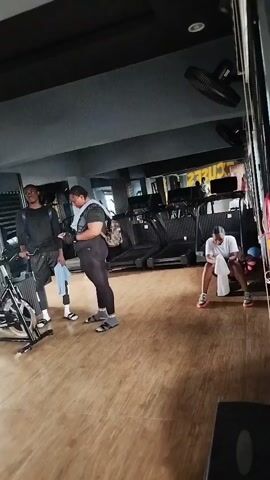 The video shows a gym with several people. In the foreground on the left, two people are standing near exercise bikes, one is holding a towel. Further back on the right, another person is sitting on a piece of equipment, also holding a towel. The gym appears to be equipped with treadmills and other exercise machines. The lighting is dim, and the overall atmosphere is one of a typical workout session.