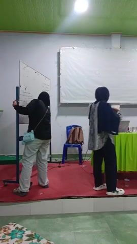 The video shows a room with a stage. On the stage, there are three people seated at a table covered with a green tablecloth. The person in the center is a man wearing a black shirt and glasses, looking at something on the table. To his right, a woman in a black headscarf and black clothing is also seated at the table, looking towards the man. To the man's left, a woman in a red headscarf and grey clothing is seated, looking towards the man as well. In front of the table, a woman wearing a black headscarf, a grey patterned shirt, and black pants is standing, facing the seated individuals. She appears to be presenting or speaking, holding a small object in her hand and gesturing with it. To the left of the stage, another person, wearing a black hoodie and light-colored pants, stands facing a whiteboard. A blue chair with a brown bag on it is visible on the stage behind the presenter. A projector screen is mounted on the wall behind the seated individuals. The room appears to be a meeting or presentation space, with a stage, a screen, and seating. The floor in front of the stage is covered in a red carpet.