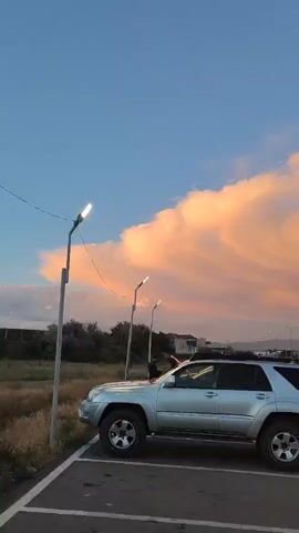 This media file captures a scenic view of a sky filled with dramatic, billowing clouds illuminated by the warm hues of sunset. The setting appears to be an urban or suburban area, likely in Ayntap, Armenia, given the context.

In the foreground, a silver SUV is parked in what seems to be a parking lot. The vehicle is facing towards the right of the frame, with its side and rear visible. To the left of the SUV, a tall lamppost with a light fixture is present, suggesting the presence of a public area or street. Further back, several other lampposts are visible, as well as a billboard structure, empty and awaiting advertisement.

The main focus of the media is the sky. The clouds are a dominant feature, painted in shades of pink, orange, and yellow, indicating a sunset or sunrise. The clouds themselves are textured and layered, creating a visually striking natural display. The sky transitions from a clear, light blue at the top to the warmer tones of the sunset clouds as it moves down.

In the background, a low-rise cityscape is visible, with buildings and trees lining the horizon. A road or highway is also present, with a few distant cars suggesting some level of activity. The overall atmosphere evokes a sense of calm and natural beauty, with the dramatic cloud formation adding a touch of awe. The time of day is clearly evening, due to the sunset lighting. There are no identifiable people clearly in focus, though a silhouette of a person can be seen near the SUV. No text is visible in the media file.