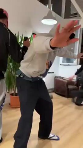 The video shows a person dancing in a living room. The dancer is wearing a grey hoodie, black pants, and blue and white flip-flops. The person is wearing sunglasses and has the hoodie pulled up over their head. Another person in a black jacket and grey pants is standing behind the dancer, holding a phone. A third person with a pink mask and a baseball cap is also in the room. The living room has a wooden floor, a potted plant, a leather chair, and a television. There's also a white ceiling with beams and a modern hanging light. The person in the hoodie appears to be rehearsing a dance routine.