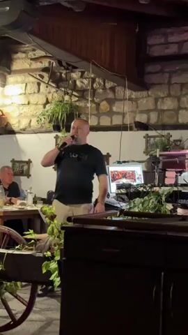 A man with short hair and a black t-shirt stands holding a microphone in his right hand. He is singing into the microphone. He is standing in front of a wooden counter. There is a computer monitor on a stand behind him with a colorful image on the screen. There are plants on the stand behind the monitor. To the left of the man, another man is seated at a table, looking towards the singer. The background features a stone wall and wooden beams, suggesting an indoor setting like a restaurant or event space.