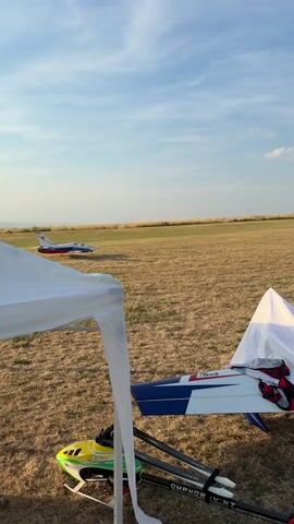 The video shows a remote-controlled jet airplane on a dry, grassy field. The airplane is white with blue and red accents and appears to be in motion, possibly taxiing. In the foreground, parts of a white canopy or tent are visible, along with a remote-controlled helicopter. The setting is an open field, likely an airfield or RC flying site. In the background, there are power lines, a communication tower, and a wind turbine, suggesting a rural or semi-rural location. A yellow marker with "1/2" is visible on the field. The sky is blue with some scattered clouds, and the lighting suggests it is late afternoon or early evening. The overall mood is peaceful and suggests an outdoor hobby activity. There is no visible text on the remote-controlled jet, but the helicopter has text that reads "OMPHOB M7".