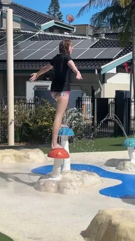A person is standing on a mushroom-shaped water spray toy in a splash pad. The person is wearing a black t-shirt and patterned shorts. Water is spraying from the mushroom and from other spray toys in the background. The ground is made of sand and blue artificial turf. Buildings with solar panels are visible in the background. The sky is blue and clear.