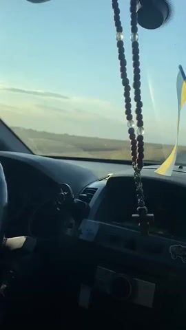The video shows the interior of a car from the passenger's perspective. A rosary hangs from the rearview mirror. The Ukrainian flag is visible on the dashboard. A hand is on the steering wheel. The car appears to be on a road in a rural area. The sky is clear and blue, with some clouds. The sun is setting.