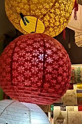 The video displays a close-up shot of several hanging lanterns. The foremost lantern is large and red, adorned with intricate black dragon designs. Directly above it is a round, red lantern patterned with smaller white floral motifs. Further up, a yellow lantern with a similar floral pattern is visible. At the bottom of the frame, a white lantern decorated with red butterflies is partially visible. The background appears to be a shop or market stall, with more lanterns and displayed items suggested by the blurred elements. The lighting suggests an indoor setting.