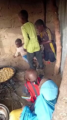 The video shows children gathered around a cooking area in what appears to be a simple dwelling with brick walls and a rudimentary roof. One child is cooking small, round items in a large pan over a fire. Another child stands nearby, looking at the pan, holding a bowl of batter. Other children are playing and moving around in the background. The environment appears modest, suggesting a low-income setting. There are metal bowls and other cooking utensils visible in the cooking area.