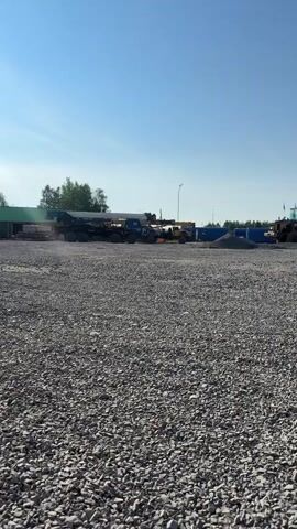 A wide shot captures a clear blue sky above a gravel lot. In the background, a large construction crane is parked, its boom extended. To its right, a yellow front-end loader sits idle. Further back, a pile of gravel is visible, and a blue tarp covers an object. The scene suggests a construction site or storage yard on a sunny day.