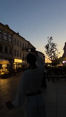 A person is filmed from behind while dancing in a street at dusk. The person is wearing a white cropped sweater and white pants. They are holding a phone in their right hand, which is raised above their head, and appear to be recording themselves. Their left arm is extended to the side. The background shows buildings lining the street with lights from shops and restaurants. A tree is visible in the middle ground. The sky is a gradient of dark blue to orange.