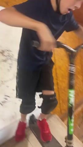 A child, wearing a dark t-shirt, dark shorts with white stripes, red shoes, and a helmet, rides a black scooter inside an indoor skatepark. The skatepark features wooden ramps and obstacles. The child rides the scooter towards a large ramp with a black surface, ascends it, and rides up and over it. The video captures the child's movement from various angles, emphasizing the action of riding the scooter on the ramps.