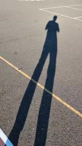 The video shows the shadow of a person standing on an asphalt surface with yellow and white painted lines. The person's shadow is long and dark, indicating a low sun angle. The asphalt surface appears to be part of a playground or sports court, marked by the white lines. There's a small, dark object on the asphalt near the bottom of the frame. The camera is static.
