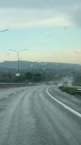 The video shows a view from inside a car driving on a highway in rainy conditions. The windshield wipers briefly clear the water droplets on the glass.  Several cars are visible on the road ahead. There are streetlights lining the side of the road and green hills in the distance. The sky is overcast.